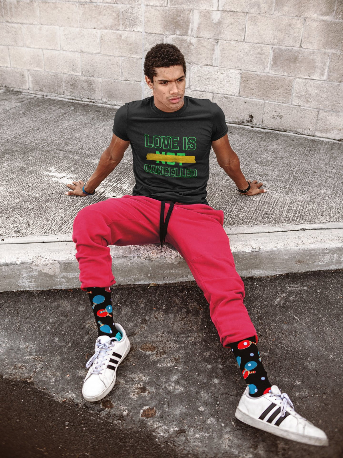 Person sitting on a concrete ledge wearing a black t-shirt with text, red pants, and colorful socks.