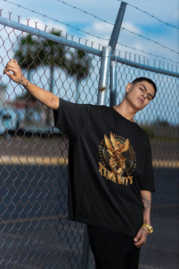 Person wearing a black t-shirt with a gold eagle design, standing against a chain-link fence.