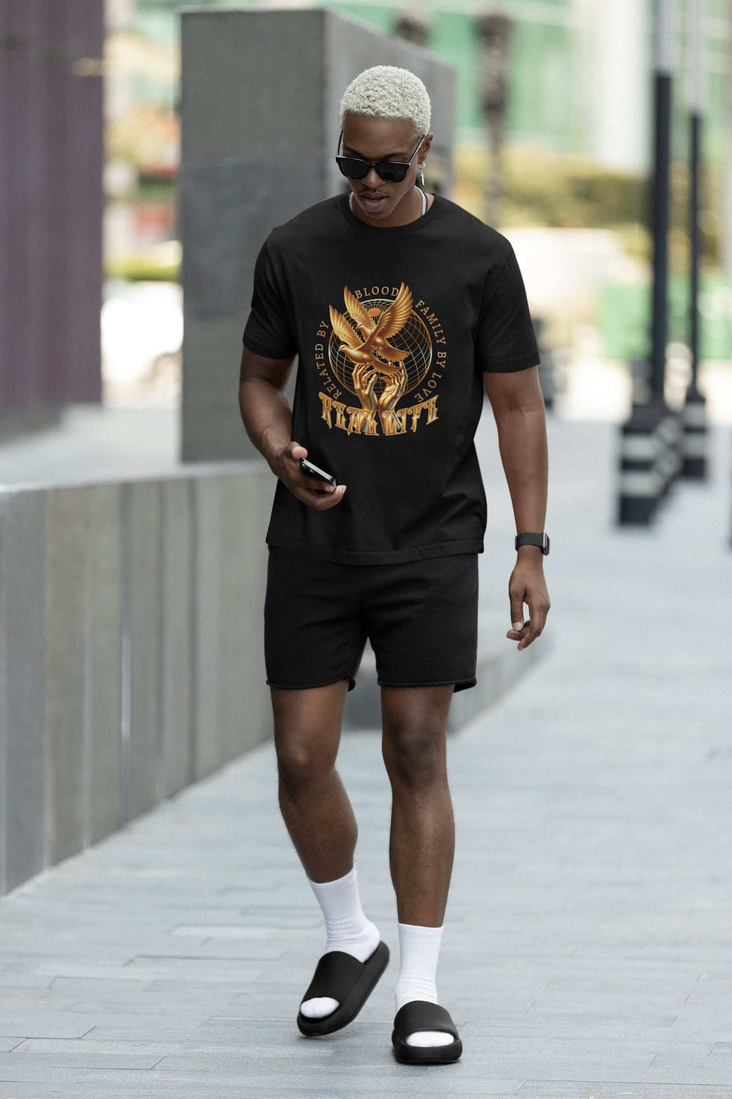 Man walking outdoors wearing a black t-shirt with a gold graphic and text, black shorts, white socks, and black sandals.
