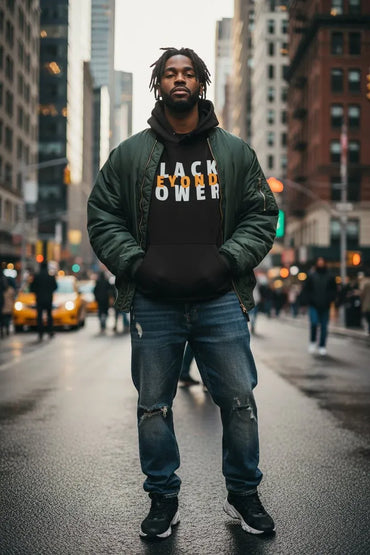 A man stands on a city street wearing a dark green puffer jacket over a black hoodie with the words 'LACK FUTURE POWER' in white and orange, paired with distressed blue jeans and black sneakers.