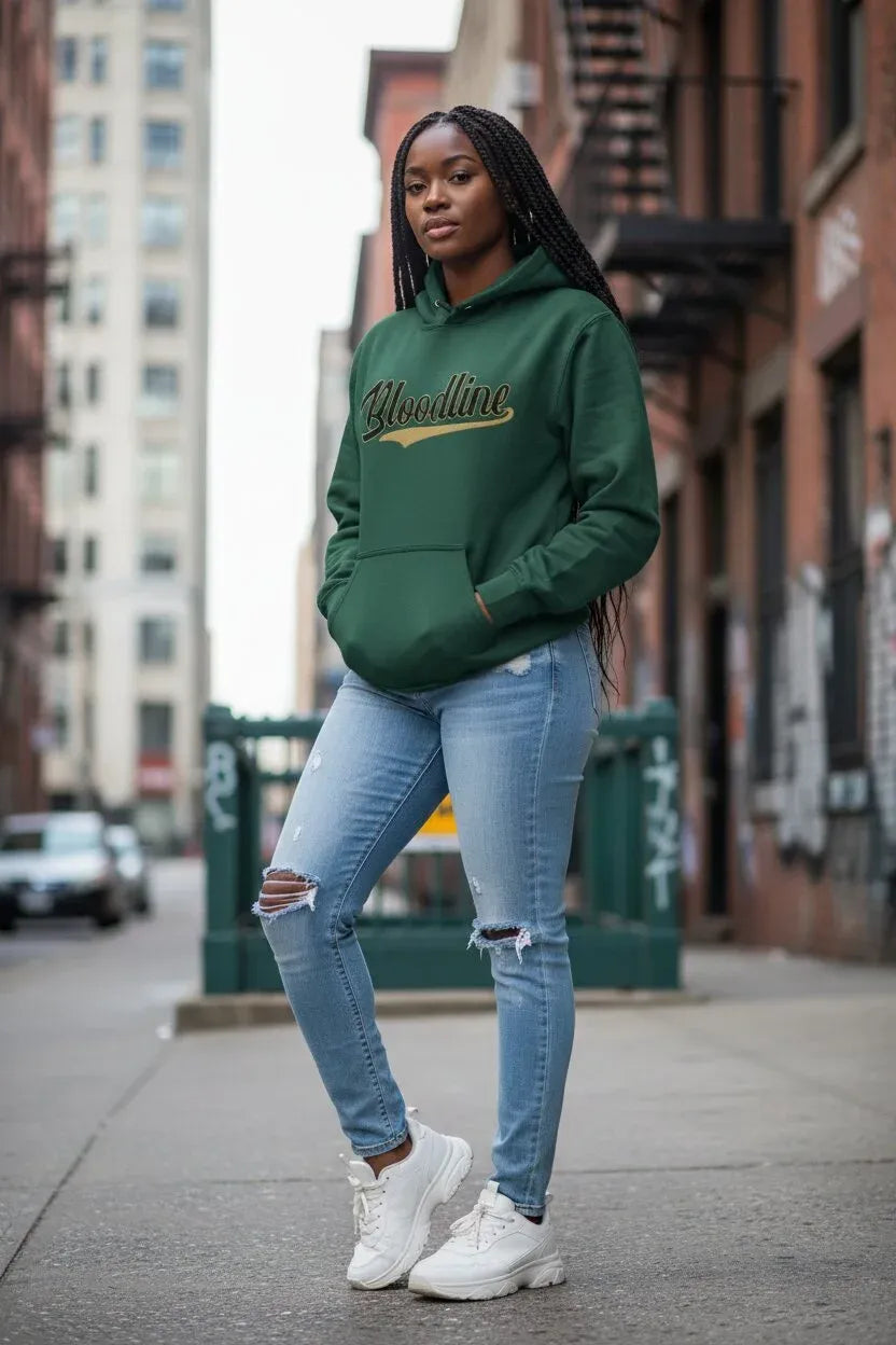 A woman stands on a city sidewalk wearing a dark green pullover hoodie with 'Blindline' written in gold script across the chest, paired with light-wash ripped jeans and white sneakers.