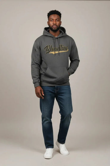 A man modeling a dark gray pullover hoodie with 'Bloodline' printed in gold script across the chest, paired with dark blue jeans and white sneakers, against a neutral studio backdrop.