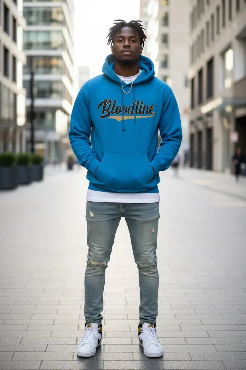A man standing on a city sidewalk wearing a bright blue hoodie with 'Bloodline' printed across the chest in black and gold, paired with distressed gray jeans and white sneakers.