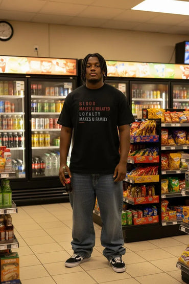A man wearing a black graphic t-shirt with red and white text, blue jeans, and black sneakers stands inside a convenience store aisle with refrigerated beverage displays and snack shelves in the background.