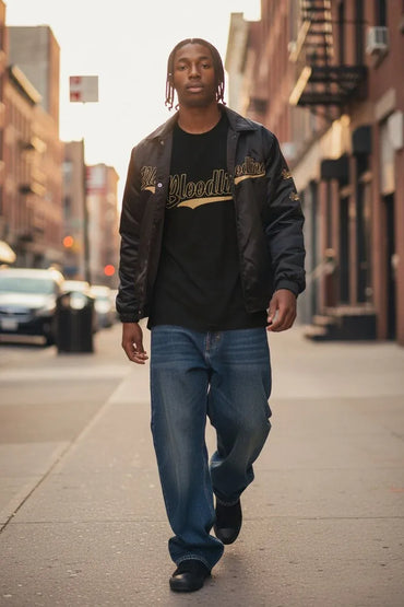 A man walking down a city street wearing a black bomber jacket with gold lettering, a black graphic t-shirt, and blue jeans.