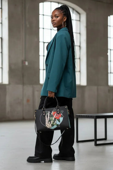 A woman in a teal blazer and black pants stands in a concrete industrial loft, holding a black leather handbag with a skeleton hand graphic and a red rose detail.