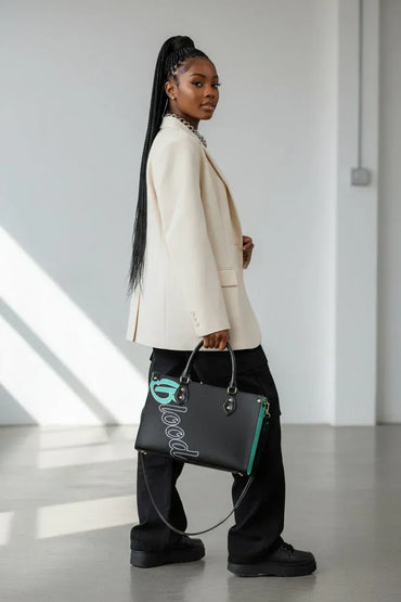 A woman in a cream blazer and black pants carries a black leather tote bag with turquoise and white script lettering in a minimalist white studio setting.