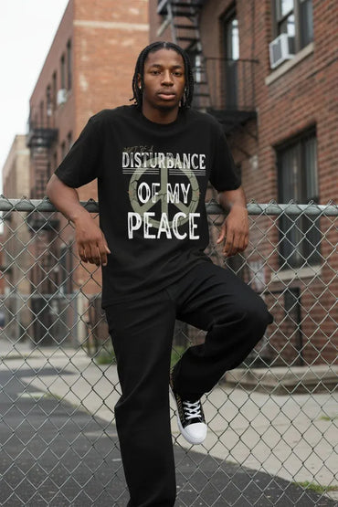 A man on a city street fence wearing a black graphic t-shirt with the text 'DISTURBANCE OF MY PEACE'