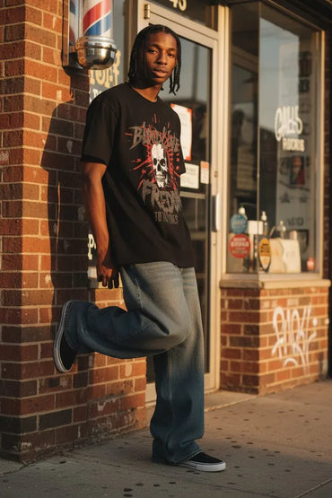 A man stands on a city sidewalk in front of a brick storefront, wearing a black graphic T-shirt with a skull print, loose blue jeans, and black sneakers.