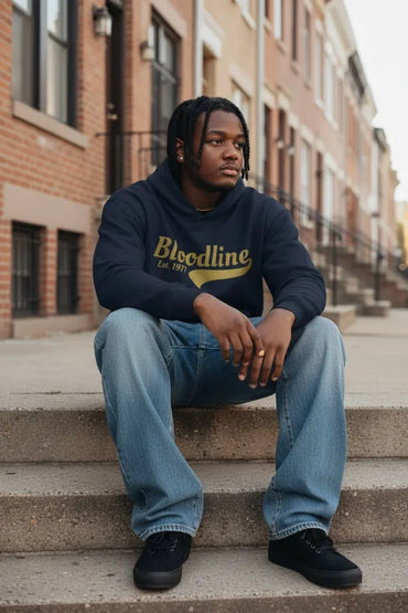 A man sitting on concrete steps in front of brick townhouses, wearing a navy blue hoodie with 'Bloodline Est. 1975' printed in yellow across the chest, paired with light blue jeans and black sneakers.