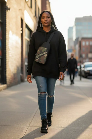 A woman walking on a city sidewalk wearing a black hoodie, ripped blue jeans, black boots, and a camouflage-patterned crossbody bag.
