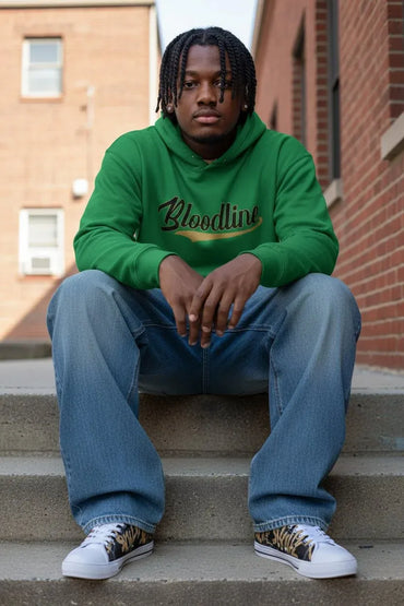 A young man with braided hair sits on concrete steps between brick buildings, wearing a bright green hoodie with 'Bloodline' printed across the chest in black script with a gold outline, blue jeans, and white low-top sneakers with a leopard print pattern.