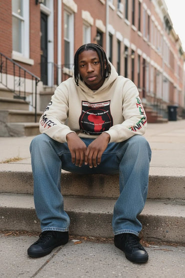A man sits on concrete steps in front of brick townhouses, wearing a cream-colored hoodie with a red and black graphic print on the front, blue jeans, and black sneakers.