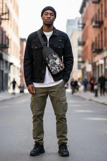 A man stands on a city street wearing a black denim jacket, black beanie, white t-shirt, olive green cargo pants, and black sneakers, with a black crossbody bag featuring a graphic print across his chest.