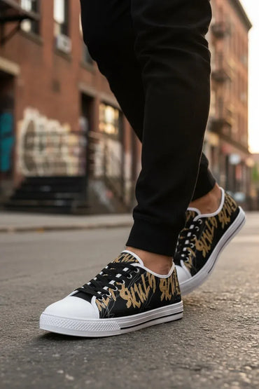 A person wearing black low-top sneakers with a gold abstract pattern on the sides, black pants, and white rubber soles, standing on a city street with blurred brick buildings in the background.