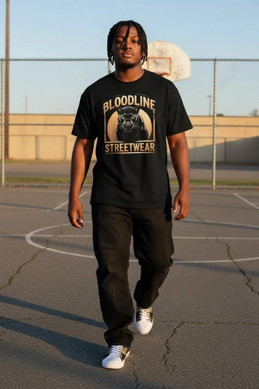 A man walking on an outdoor basketball court wearing a black graphic t-shirt with 'BLOODLINE STREETWEAR' and a panther design, paired with black sweatpants and white sneakers.