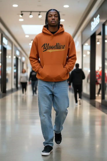 A man walking down a mall corridor wearing an orange hoodie with 'Bloodline' printed across the chest in black and gold, paired with light blue jeans and black sneakers.