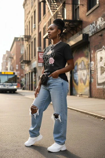 A woman stands on a city street wearing a black graphic T-shirt with a pink rose design, light blue ripped jeans, and white sneakers. Brick buildings with graffiti and a bus are visible in the background.