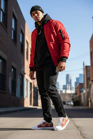 A man wearing a bright red bomber jacket, black hoodie, black pants, and red-and-white patterned sneakers, standing on an urban street.