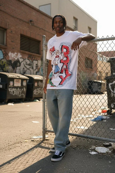 A man wearing a white graphic t-shirt with a colorful 'BAY' design, light blue jeans, and black sneakers, standing outdoors next to a chain-link fence in an urban alleyway.
