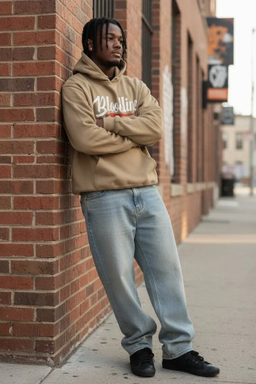 A man stands on a city sidewalk leaning against a red brick wall, wearing a beige hoodie with 'Blissline' printed across the chest, light blue jeans, and black sneakers.