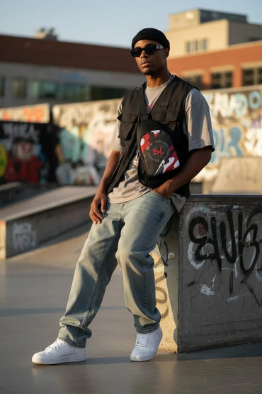 A man wearing a black beanie, sunglasses, a black utility vest over a light gray t-shirt, light blue jeans, and white sneakers, standing in an urban outdoor setting with graffiti walls in the background.