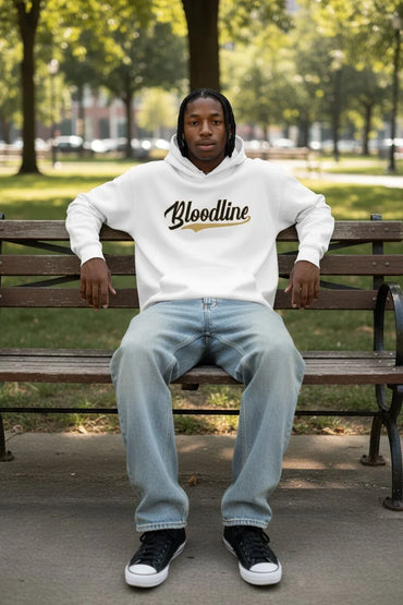 A man sitting on a wooden park bench wearing a white hoodie with 'Bloodline' printed in black and gold across the chest, paired with light blue jeans and black sneakers.