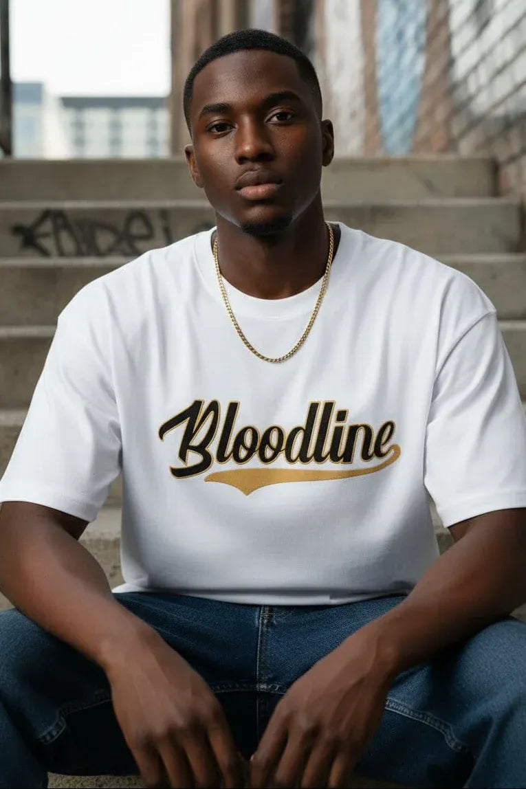 A man wearing a white crewneck t-shirt with 'Bloodline' printed in black script and gold accents across the chest, paired with blue jeans and a gold chain necklace, sitting on concrete steps.