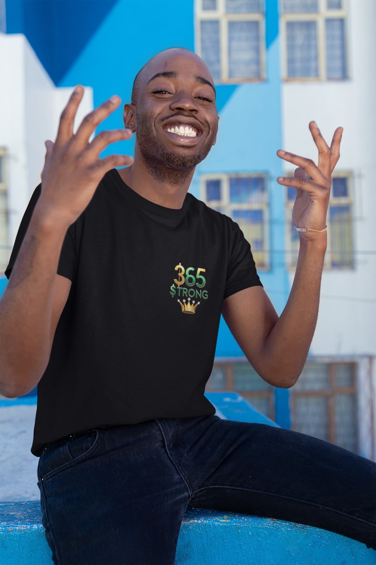 Man wearing a black t-shirt with '365 Strong' design, sitting against a blue and white building.