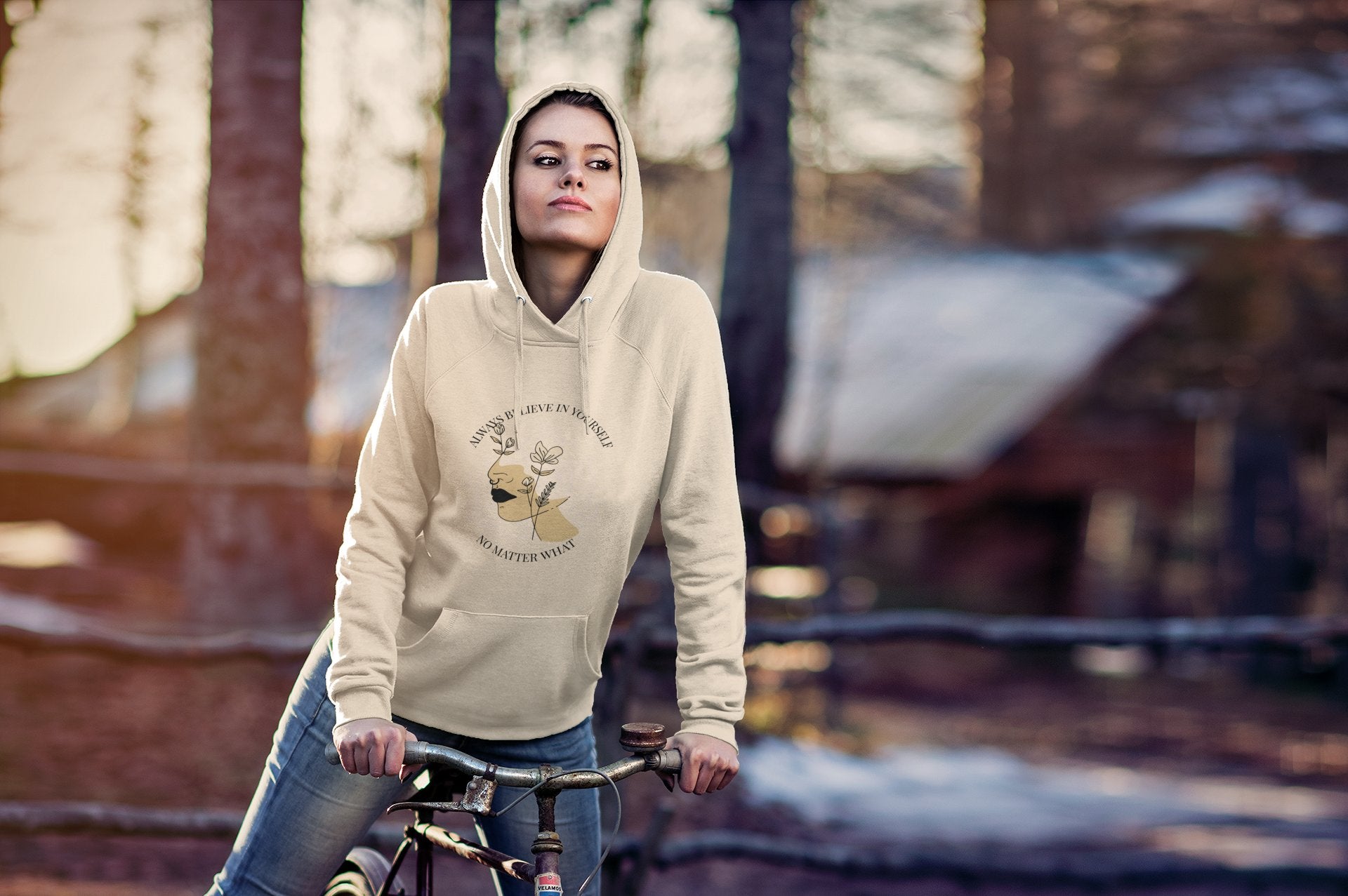Person wearing a beige hoodie with a graphic design, standing next to a bicycle in a forest setting.