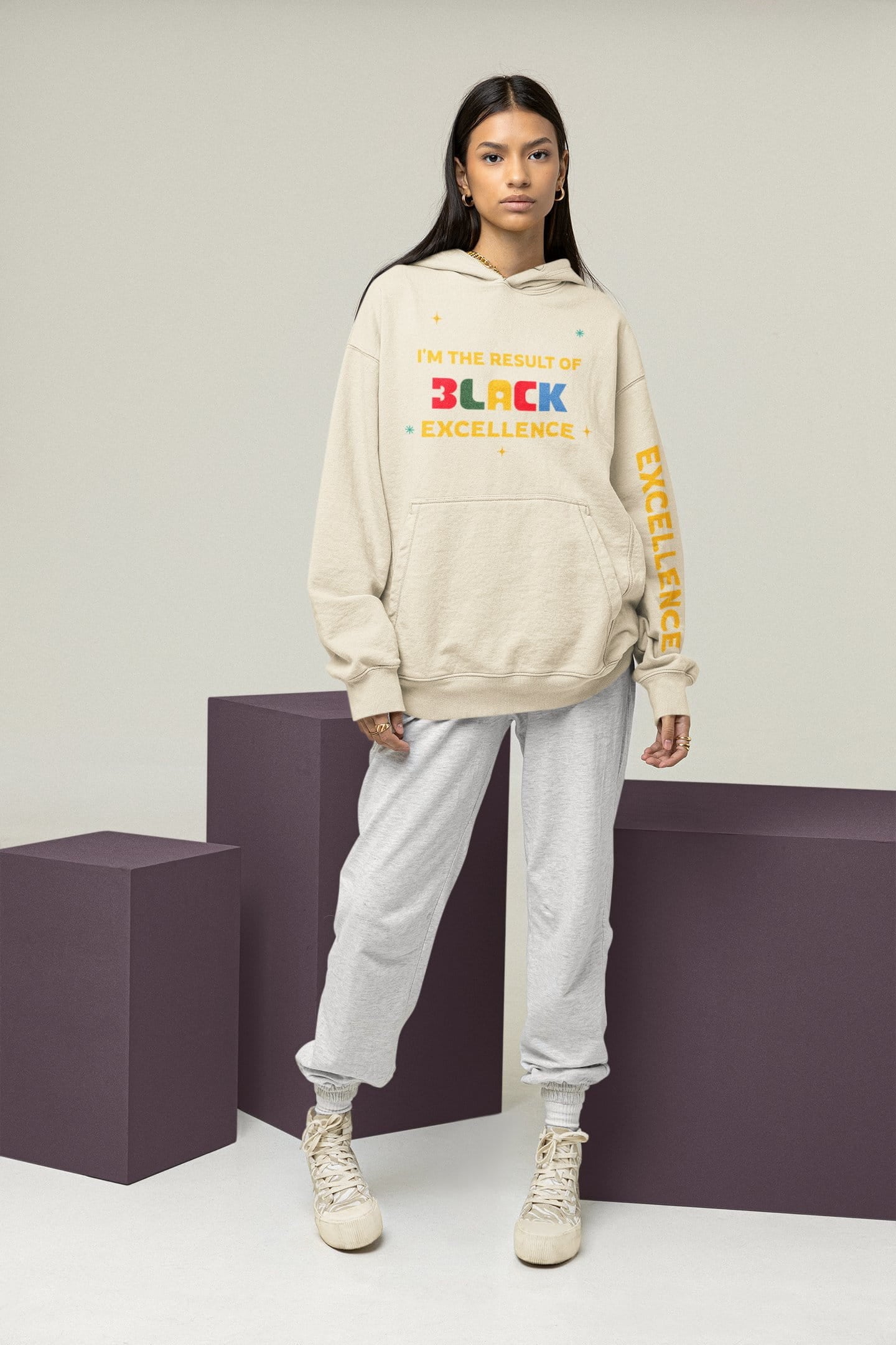 Person wearing a beige hoodie with colorful text and light gray pants on a plain background