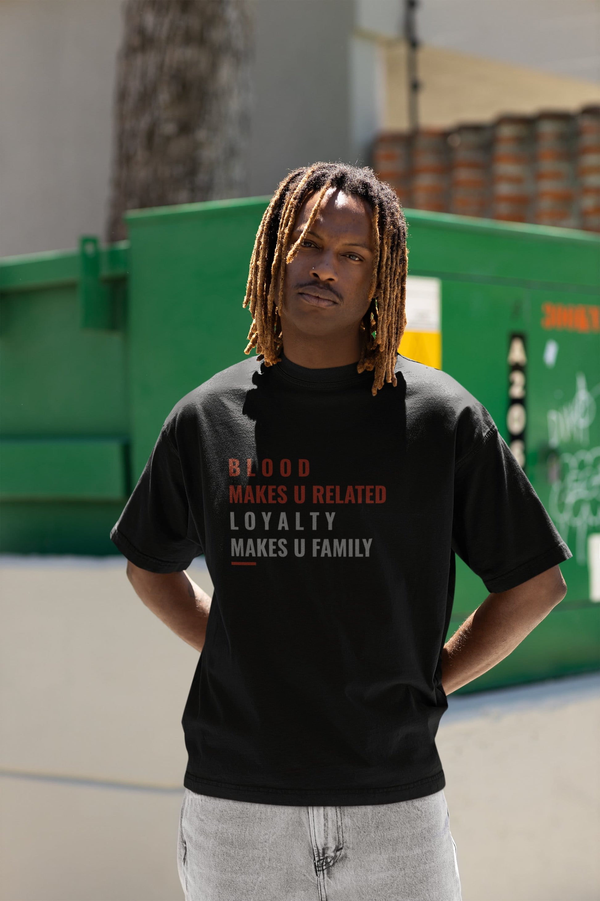 Person wearing a black t-shirt with text, standing in front of a green dumpster.