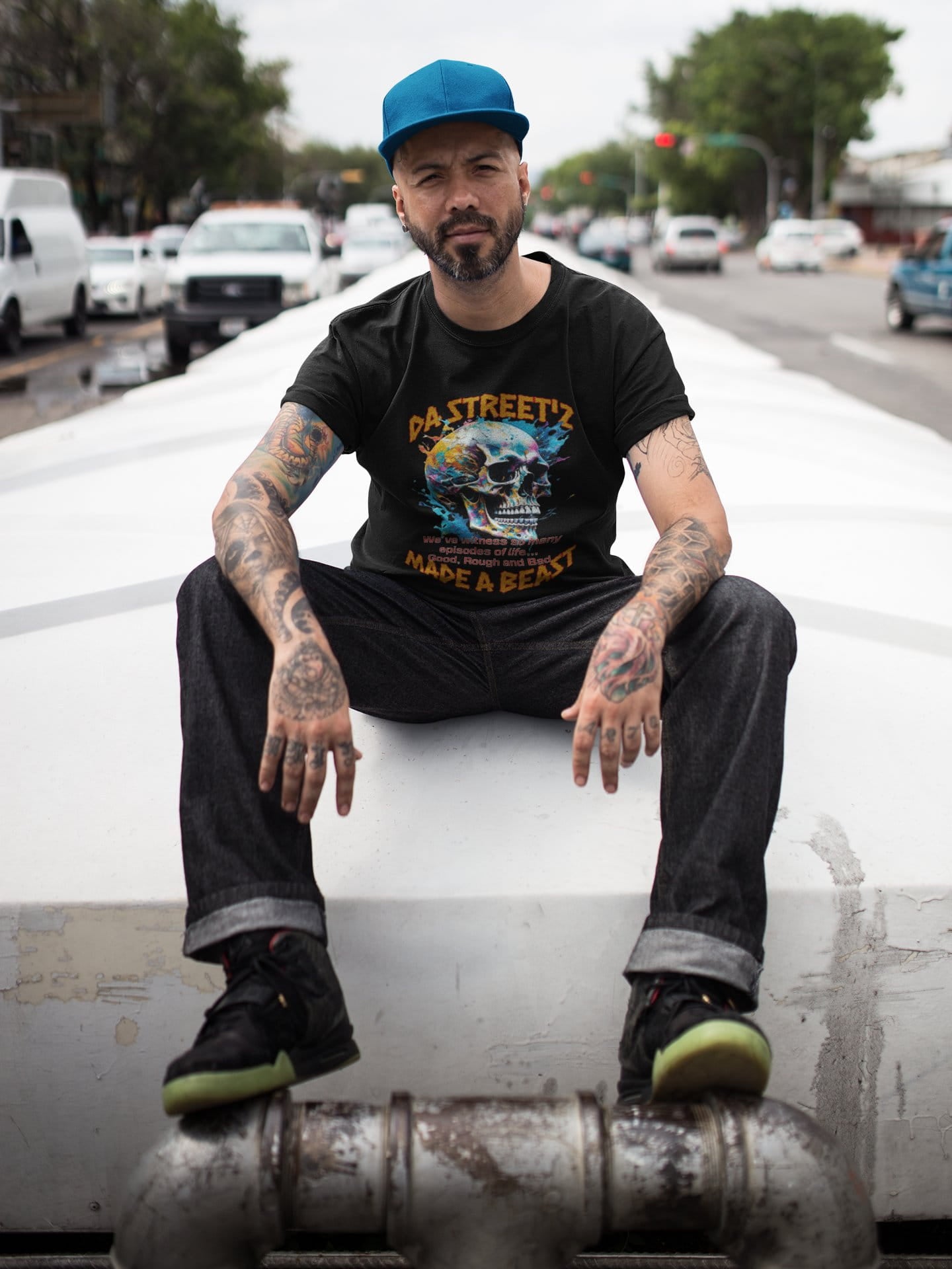 Man sitting on a car with tattoos and a beard, wearing a black t-shirt and blue cap.