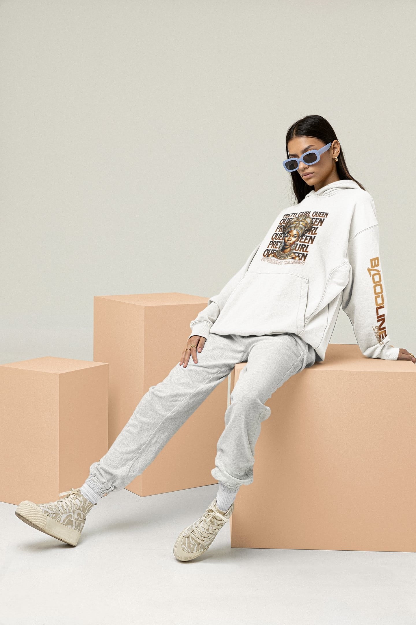 Person wearing a white hoodie with text and graphics, sitting on beige blocks against a light gray background.