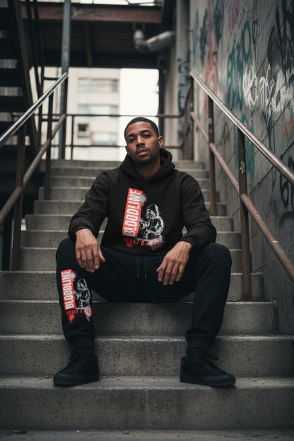 Man sitting on stairs wearing a black hoodie with graphic designs and text.