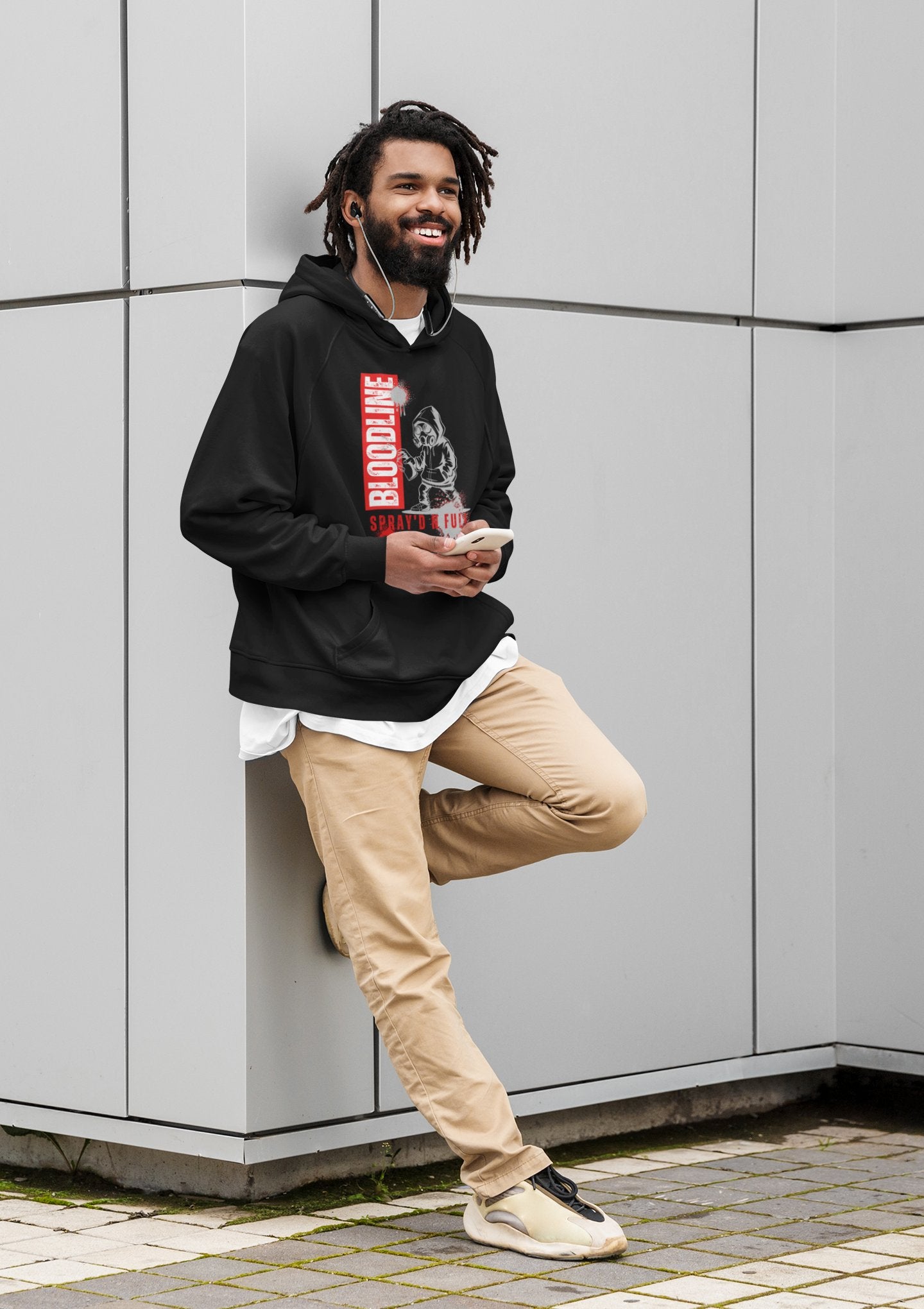 Man wearing a black hoodie with a graphic design, beige pants, and white sneakers leaning against a light gray wall.