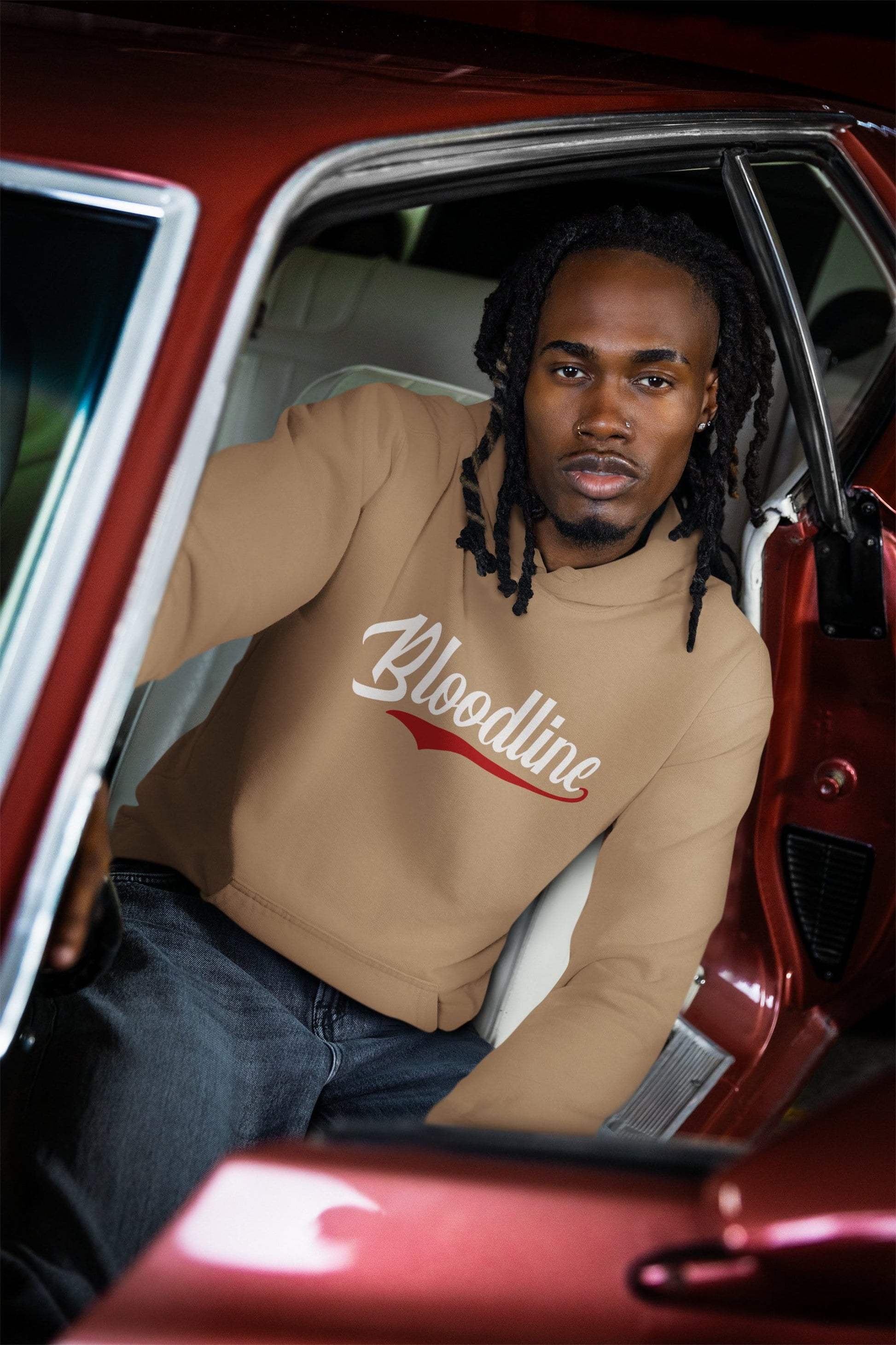 Man sitting in a red car wearing a beige sweatshirt with 'Bloodline' text.