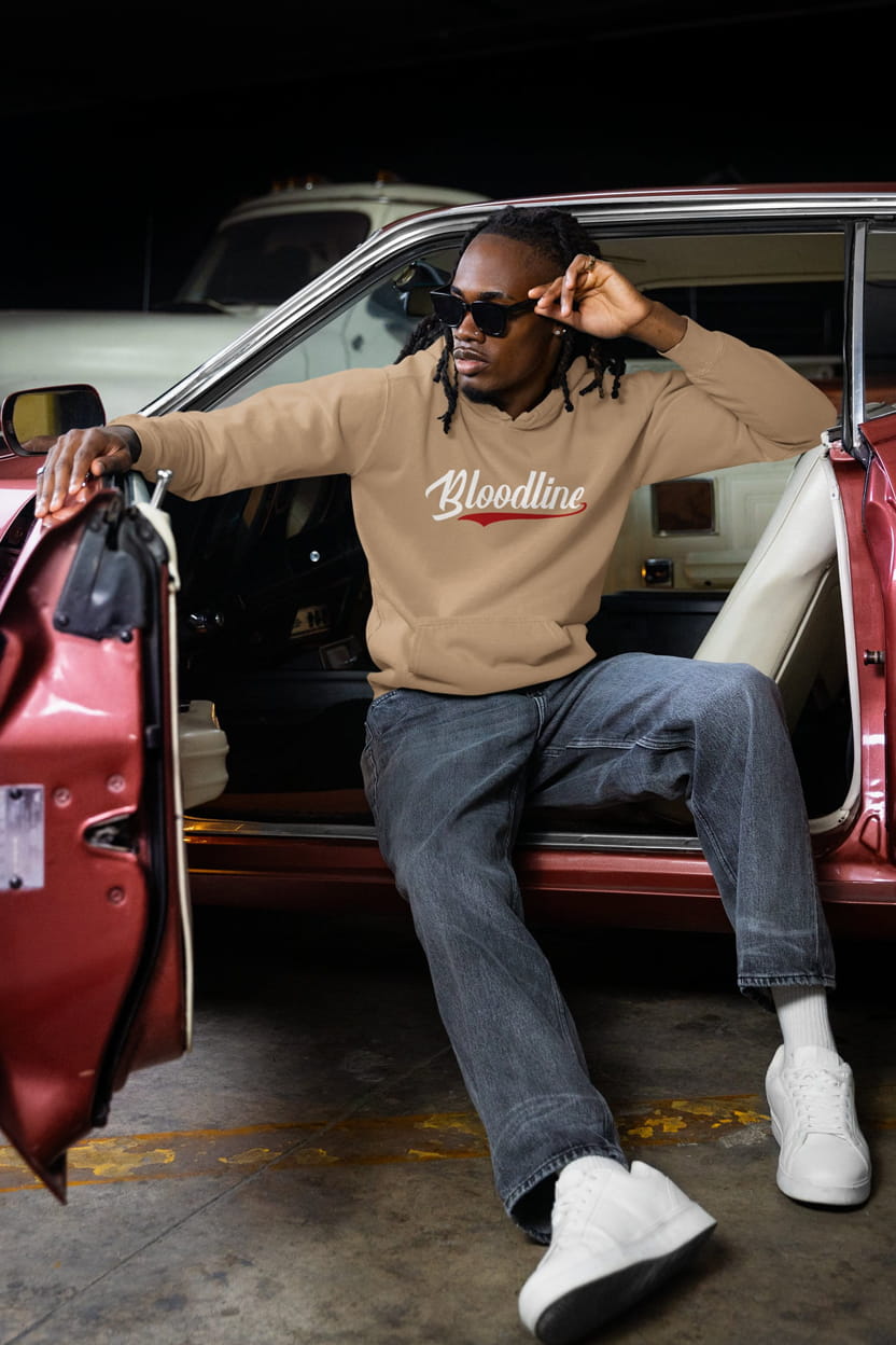 Man sitting in a car wearing a beige sweatshirt with 'Bloodline' text, sunglasses, and white sneakers.