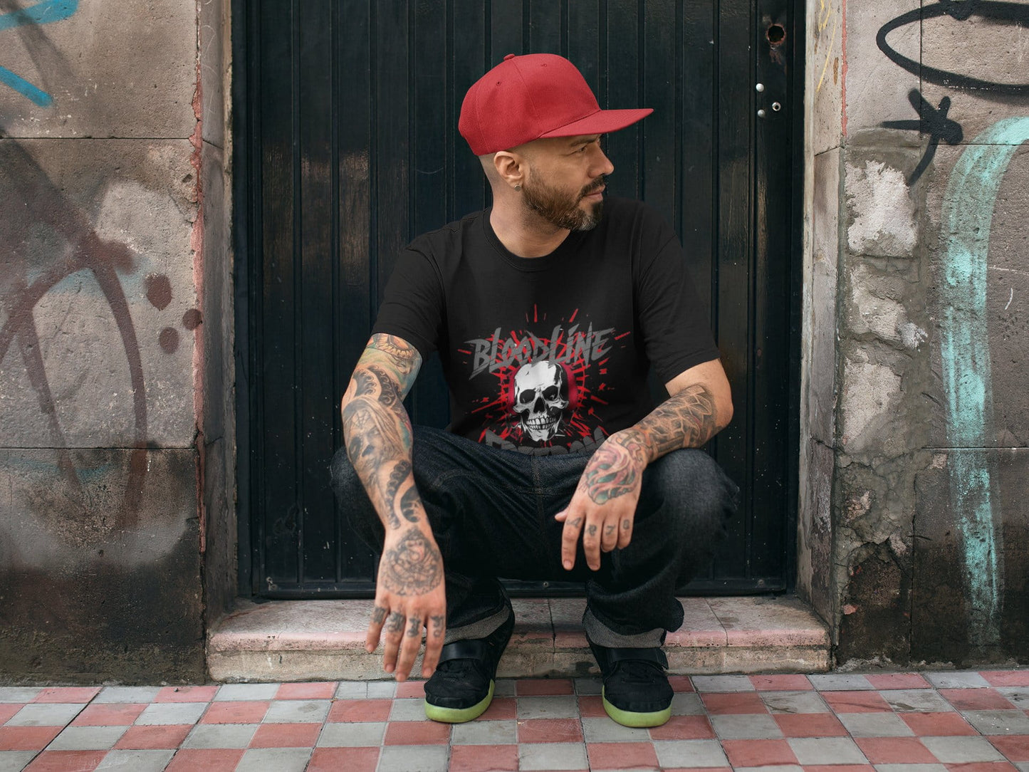 Man wearing a black t-shirt with a graphic design, sitting against a graffiti-covered wall.