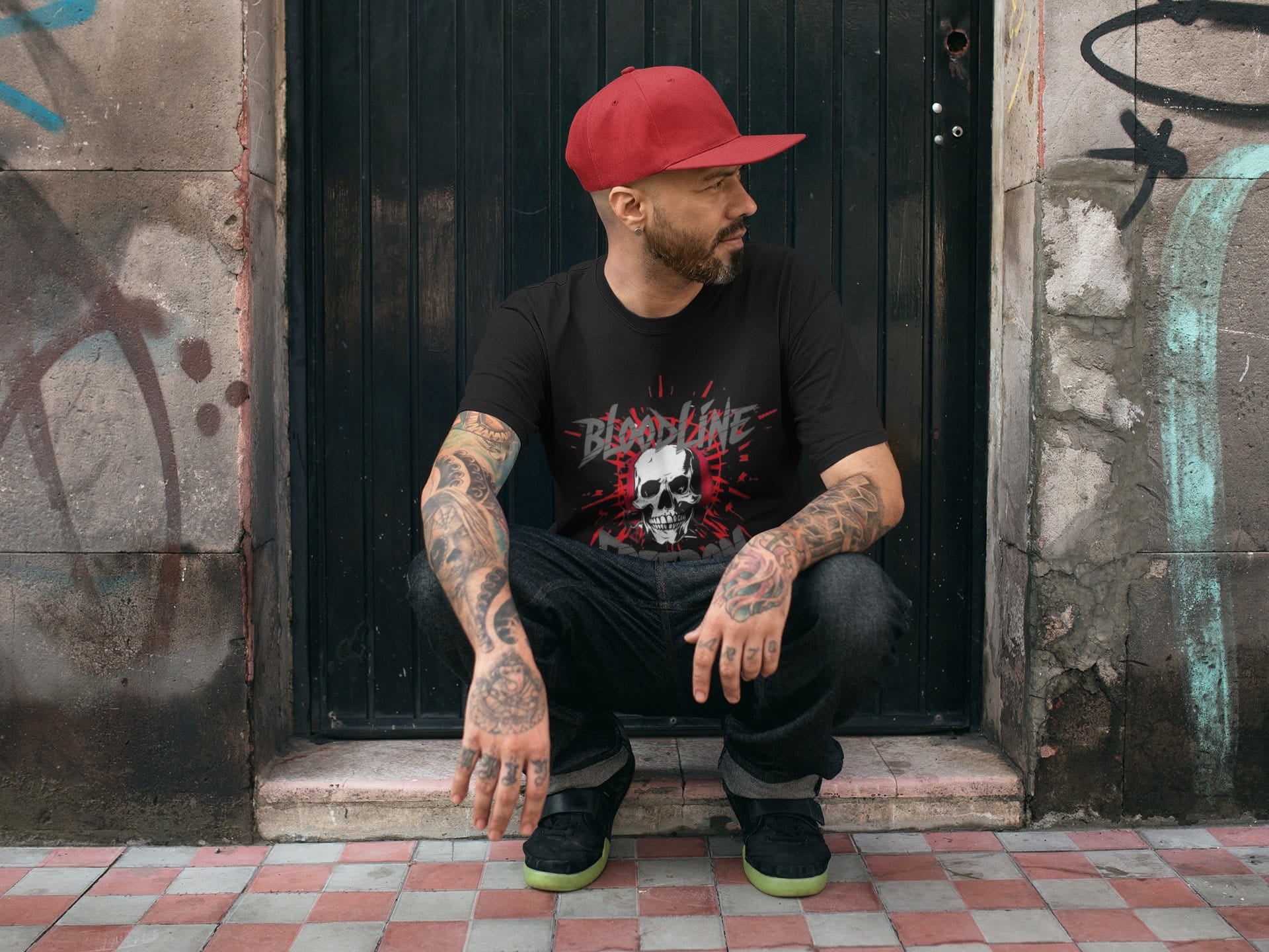 Man wearing a black t-shirt with a graphic design, sitting against a graffiti-covered wall.