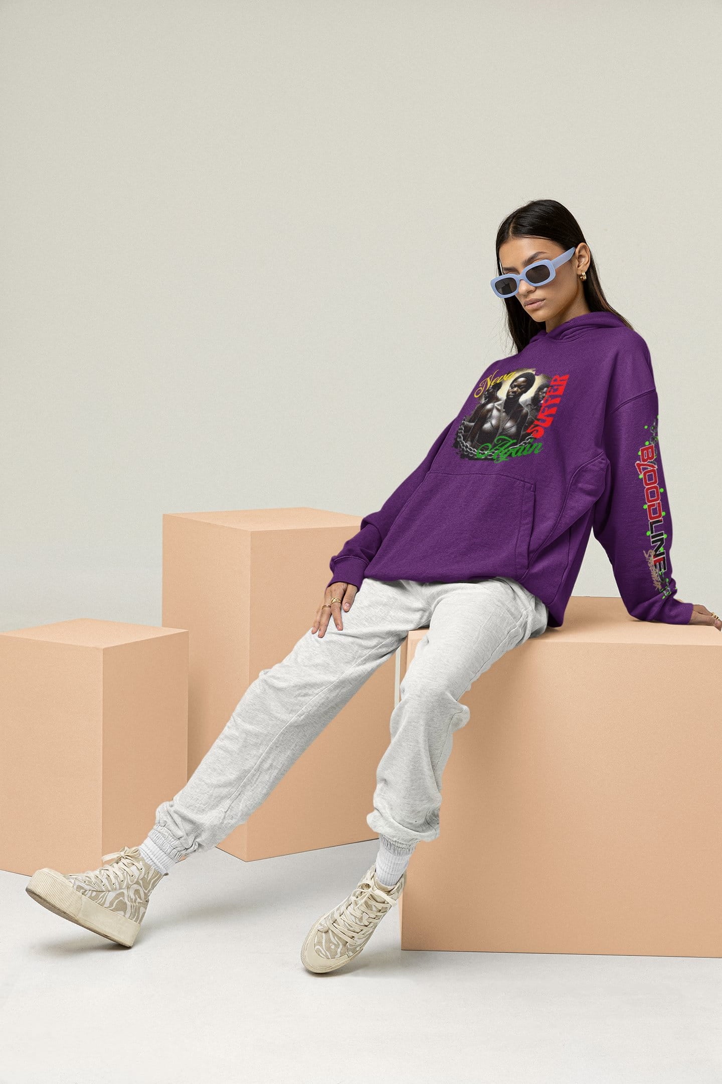 Person wearing a purple hoodie with a graphic design, sitting on a beige block against a light gray background.