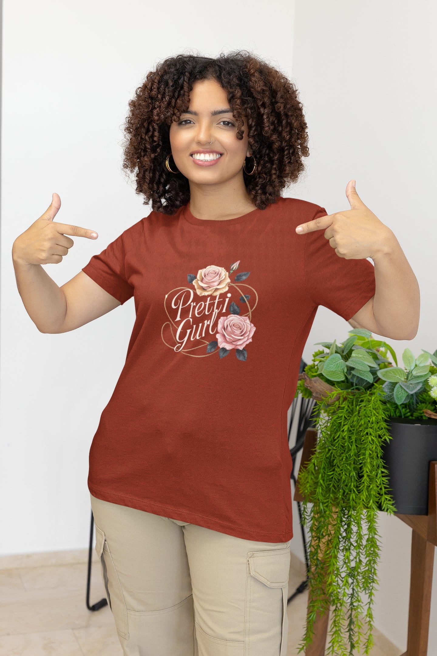 Woman wearing a red t-shirt with 'Pretty Girl' design, pointing at it, in a room with plants.
