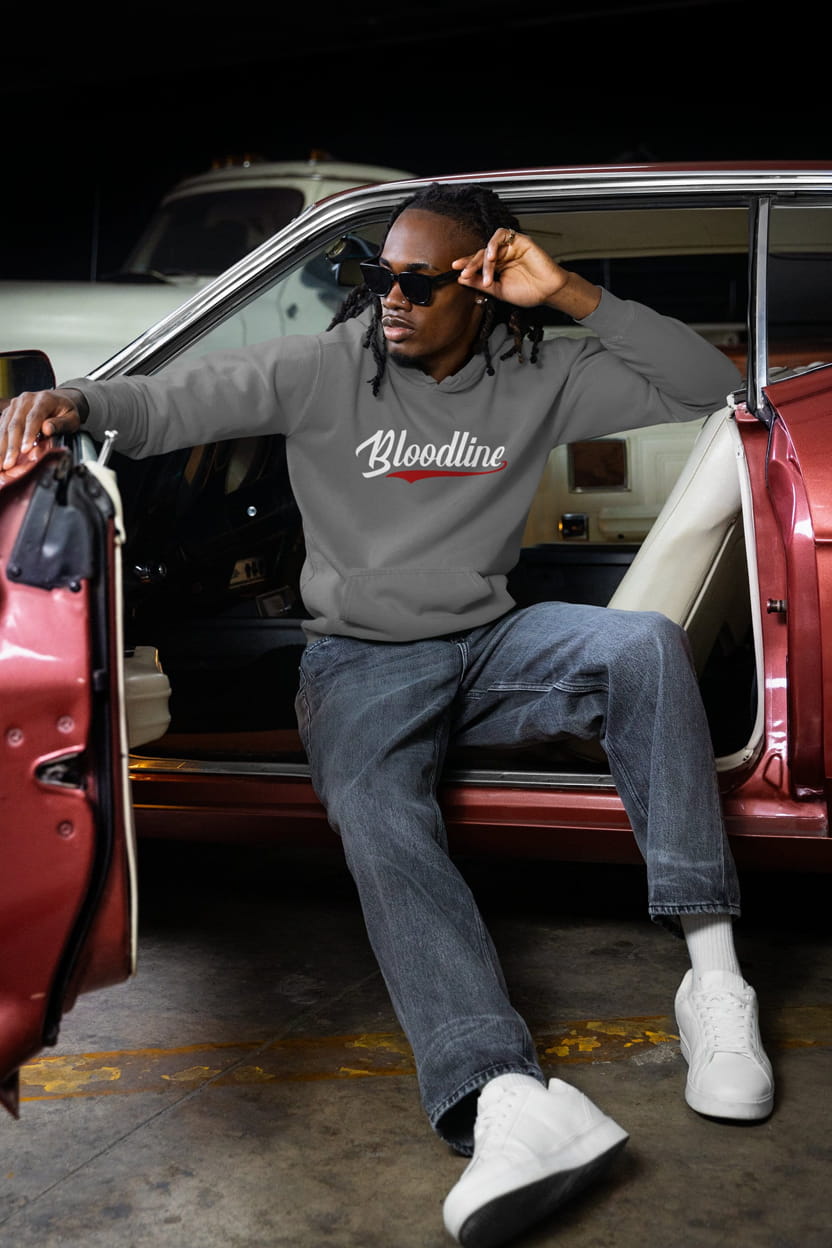 Man sitting in a red car wearing a gray sweatshirt with 'Bloodline' text.