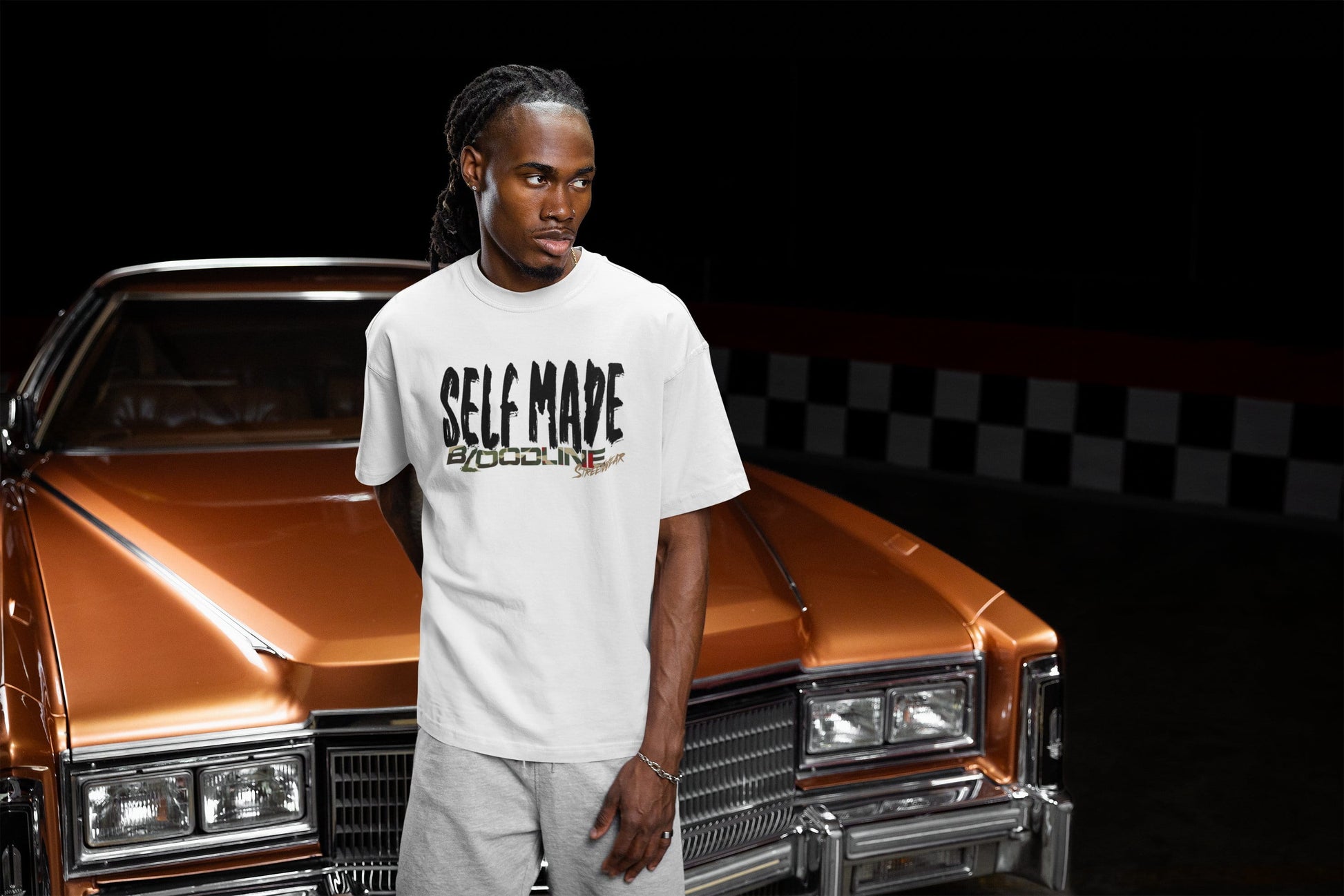 Man wearing a white t-shirt with 'SELF MADE' text standing in front of a classic car.