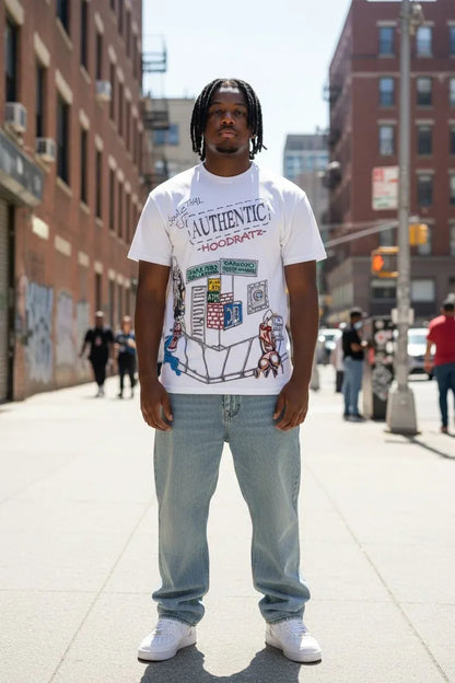 A man stands on a city sidewalk wearing a white graphic T-shirt with a colorful urban illustration and text, light blue jeans, and white sneakers. Brick buildings and blurred pedestrians are visible in the background.