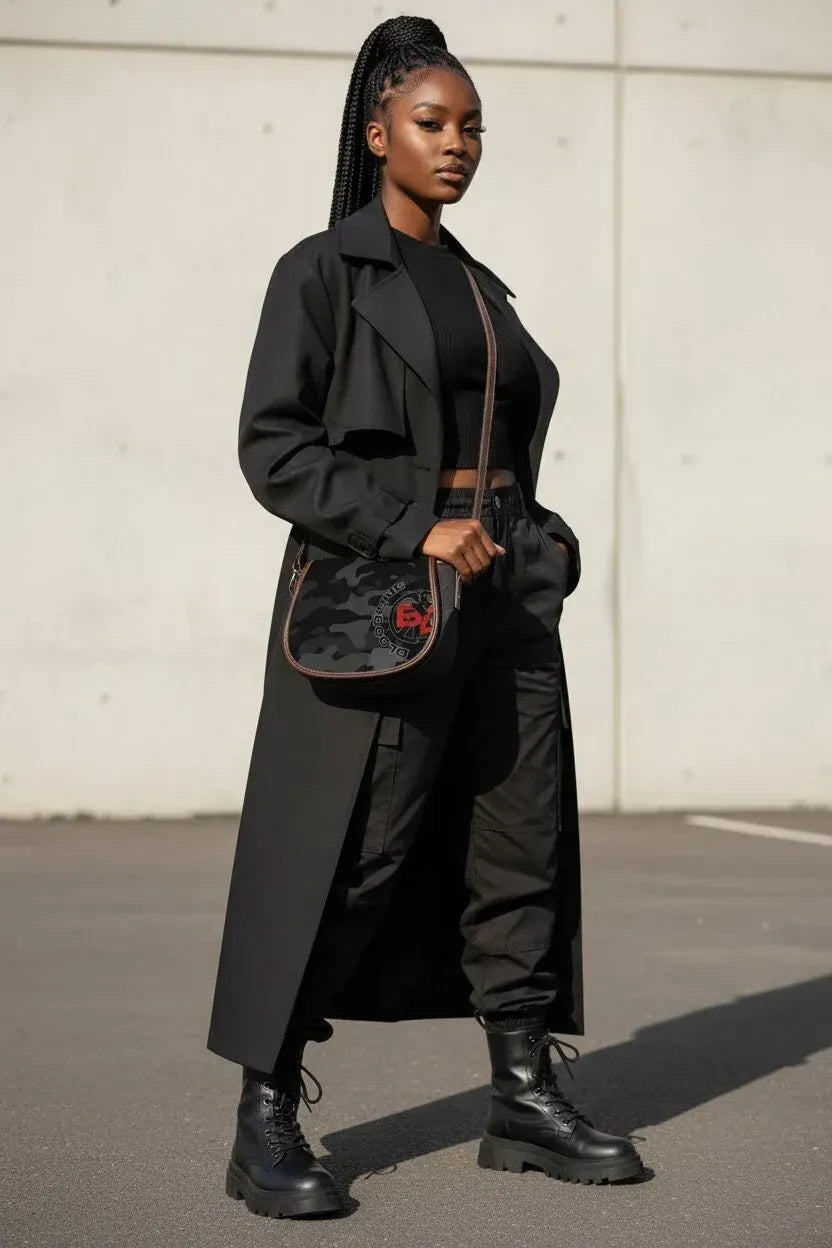 A woman stands outdoors wearing a long black trench coat over a black outfit, with black combat boots and a black crossbody bag featuring a red logo.