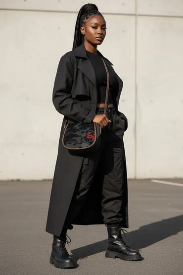 A woman stands outdoors wearing a long black trench coat over a black outfit, with black combat boots and a black crossbody bag featuring a red logo.