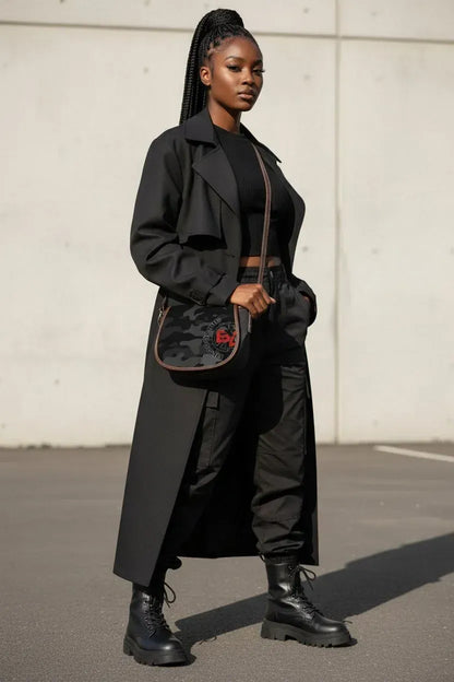 A woman stands outdoors wearing a long black trench coat over a black outfit, with black combat boots and a black crossbody bag featuring a red logo.