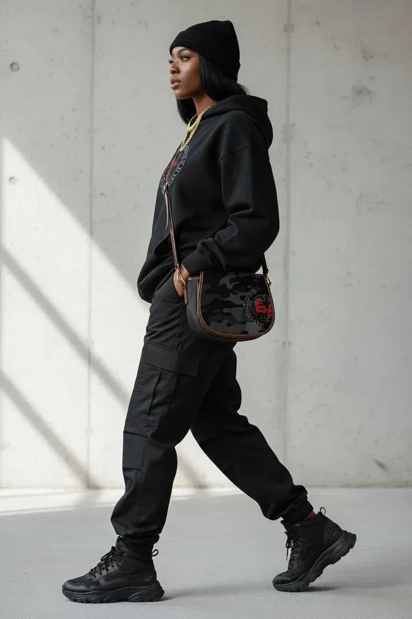 A model wearing a black beanie, black hoodie, black cargo pants, and black boots, carrying a black camouflage shoulder bag with a red rose detail, walking against a concrete wall.