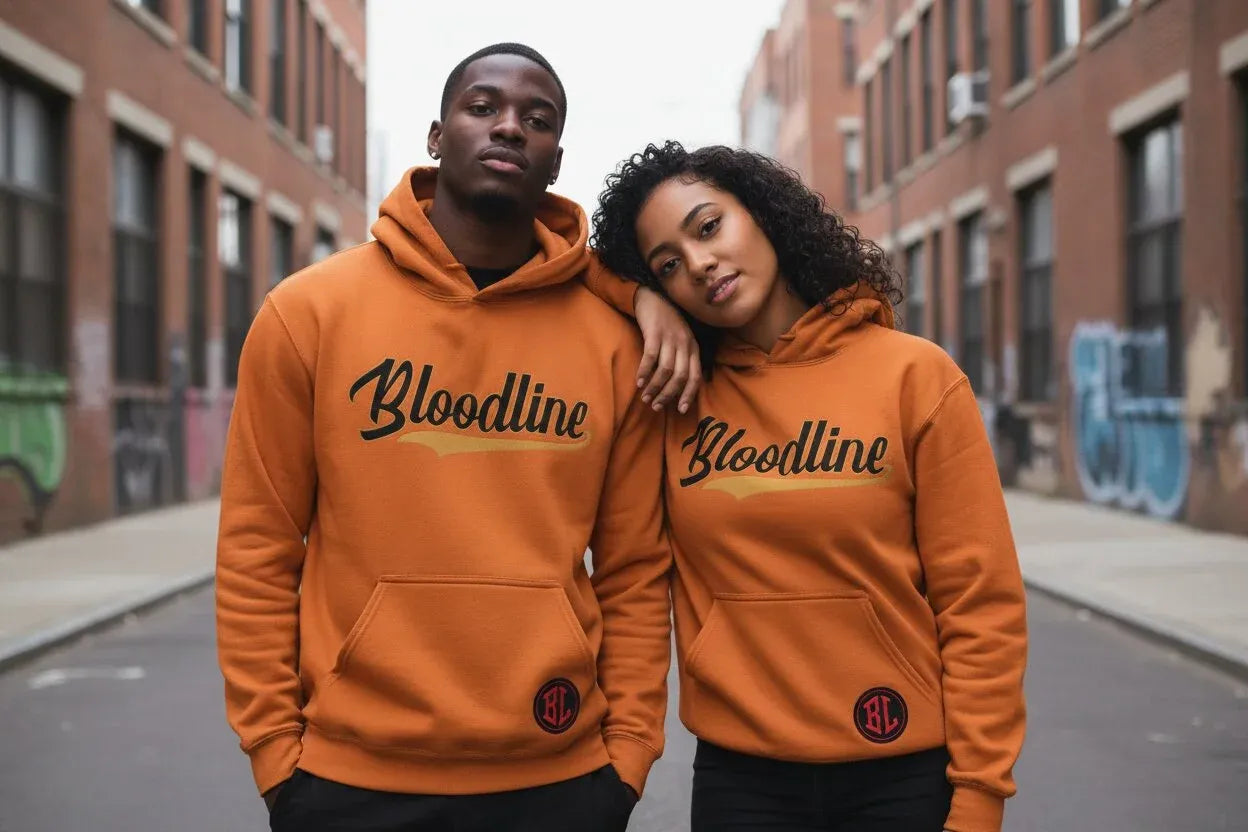 A man and a woman standing in an urban alley wearing matching burnt orange hoodies with 'Bloodline' printed across the chest in black script.